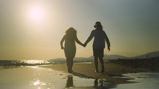 Low angle back view of young boy and girl holding hands and walking on sandy beach. Against setting sun. Summer evening at seaside.
