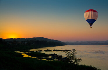 Hot air balloon flying over the Mae Kong river with scenic view of sunset in background in Northeast Thailand.