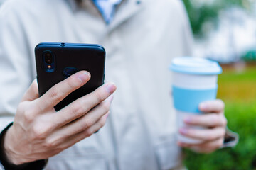 close up of a man's hand holding a smart cell phone and a portable coffee cup