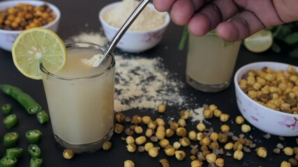 Female's hand mixing Chana powder in a glass of cool Sattu drink - tasty food. Closeup shot of a bowl of roasted Chana / chickpea  green chili  a slice of lemon against a blurred background