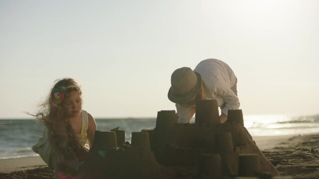 Two children playing with sand on windy beach. Siblings building sandcastle against late afternoon sun. Summer at seaside.