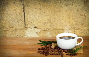  white ceramic cup of hot black americano coffee with a pile of roasted arabica coffee beans on a wooden table against a rustic concrete background. Morning refreshment drinks concept.