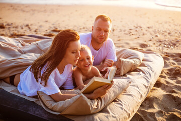 Mom is reading a book to her son while lying on the bed. Family vacation by the sea. Happy young caucasian family resting together at sunset