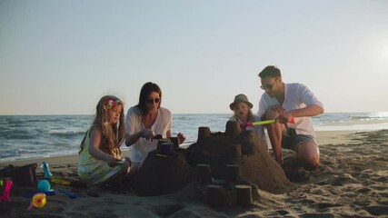 Orbit shot around parents helping children with building sandcastle on beach. Splashing waves in background. Summer at seaside.