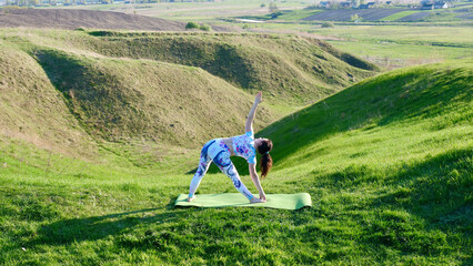 Woman yogi practicing yoga in nature, green hills in summer on a yoga mat. A brown-haired, Caucasian girl stands in an asana in sportswear, leggings and a T-shirt with braided hair in a ponytail.