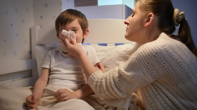 Caring mother blowing runny nose of her sick little son lying in bed. Concept of children illness, disease and parent care
