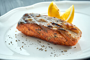 Appetizing grilled salmon steak on charcoal with lemon, served in a white plate on a dark wooden background. BBQ seafood