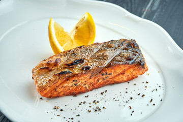 Appetizing grilled salmon steak on charcoal with lemon, served in a white plate on a dark wooden background. BBQ seafood