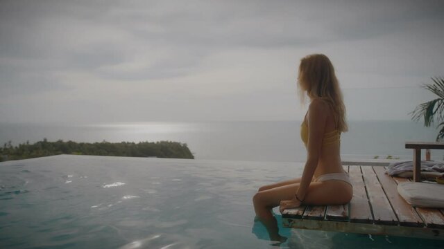 A blonde woman drinks mango shake while lounging in the sun by the pool on vacation. Elegant young woman drinks cocktail by the pool in slow motion.