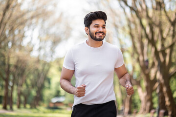 Fototapeta premium Happy muscular man dressed in sport clothes jogging during morning time at green city park. Handsome hindu guy leading active and healthy lifestyles.