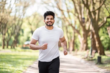 Zelfklevend Fotobehang Positive arabian man in active outfit jogging at green summer park. Young bearded guy enjoying outdoors workout. Concept of people, sport and strength. © sofiko14