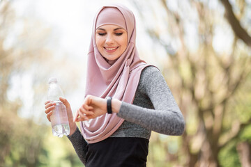 Pleasant woman in hijab looking at smart tracker for checking jogging distance. Young female athlete in sport clothing standing at green park and holding bottle of water.