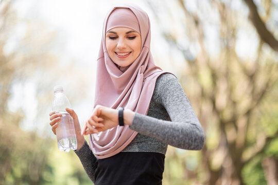 Pleasant Woman In Hijab Looking At Smart Tracker For Checking Jogging Distance. Young Female Athlete In Sport Clothing Standing At Green Park And Holding Bottle Of Water.