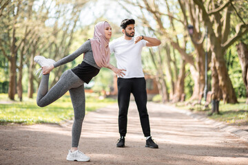 Active woman in sport clothes and hijab stretching legs at summer park. Young arabian man standing on background and using smart watch. Sport activity outdoors.