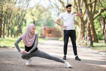 Fit young woman in hijab and sport outfit warming up at park before morning run. Handsome muslim...