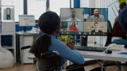Fototapeta na wymiar Paralysed handicapped black worker sitting immobilized in wheelchair having videomeeting discussing online with remotely colleagues in business office. Invalid employee working in financial company