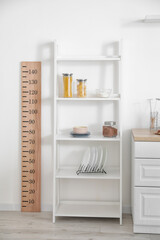 Shelves with kitchenware and stadiometer near light wall