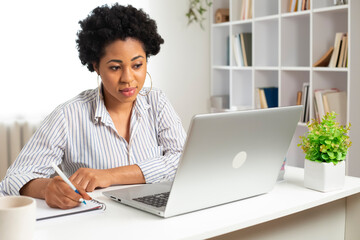 African American female student uses a laptop and writes in a notebook. Portrait of a young black woman working remotely on the computer at home. Online education.