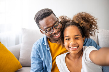 Shot of an adorable little girl taking selfies with her father at home. Smiling young dad and little daughter make selfie on smartphone. 