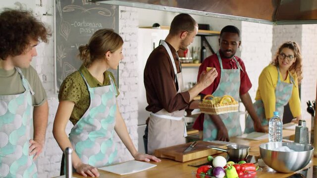 Male chef giving tagliatelle pasta to young multiethnic people and explaining recipe during culinary master class in kitchen