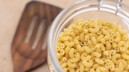 The uncooked macaroni in the opened bottle with the blurred wooden turner in the background