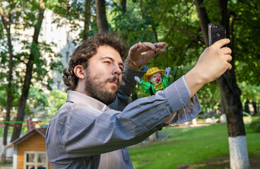 A young dark-haired man with a beard with a clown toy in his hand takes pictures of himself on a mobile phone on a natural background