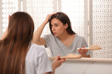 Young woman with hair loss problem looking in mirror indoors
