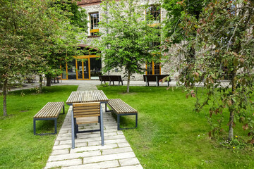Empty place for rest in a park (wooden table and benches) on a summer day. Green trees and stone paving. Brick building facade with big glass windows and door on the back. Ulemiste, Tallinn, Estonia