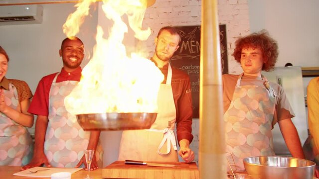 Zoom In Of Male Chef Adding Alcohol To Pan And Flambeing Food During Cooking Master Class As Group Of Multiethnic Students Watching Flames With Excitement