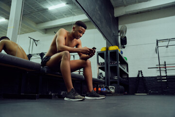 Fototapeta na wymiar Young African American male looking at his phone to reply to an online message. Mixed race, male personal trainer looking at his phone while sitting down in the gym. High quality photo 