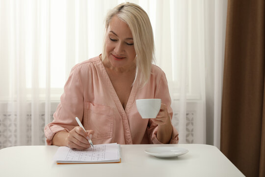 Middle aged woman with cup of drink solving sudoku puzzle at table indoors
