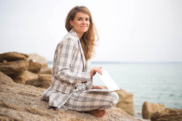 young woman sits on a stone by the sea with a laptop. remote work concept
