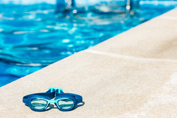 Blue swimming goggles on the curb of the pool, in the lower left corner, with the blue water in the...