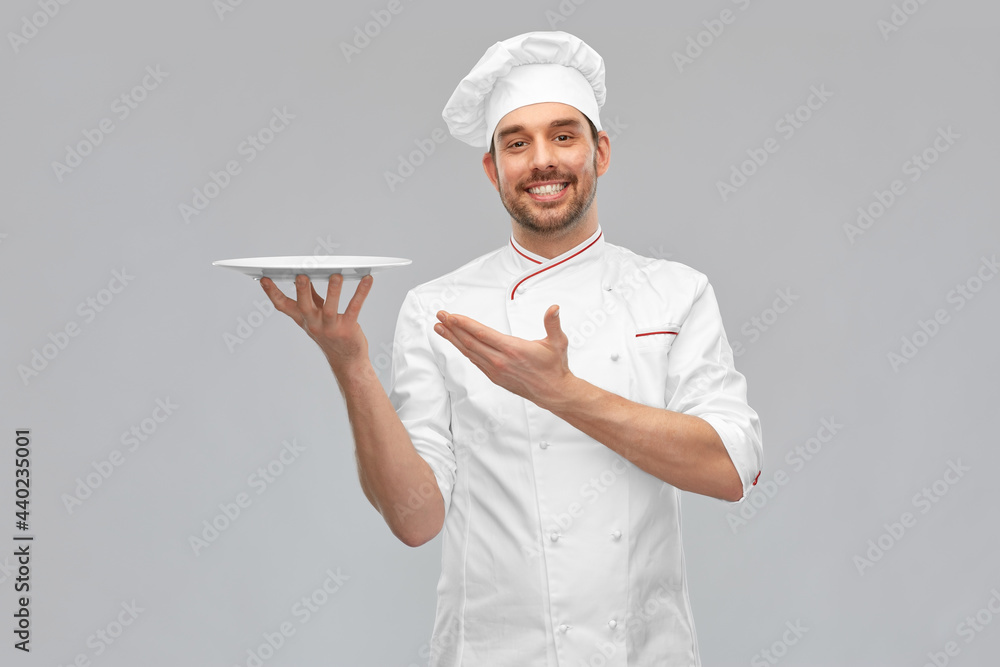 Wall mural cooking, culinary and people concept - happy smiling male chef in toque holding empty plate over grey background