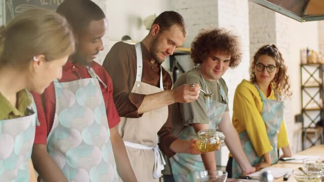 Male chef adding honey to bowl and explaining recipe to diverse group of young men and women while giving cooking master class