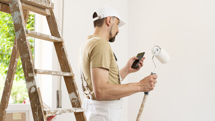 house painter man with mobile phone and paint roller, works the white home wall, a wooden ladder and green window as a background