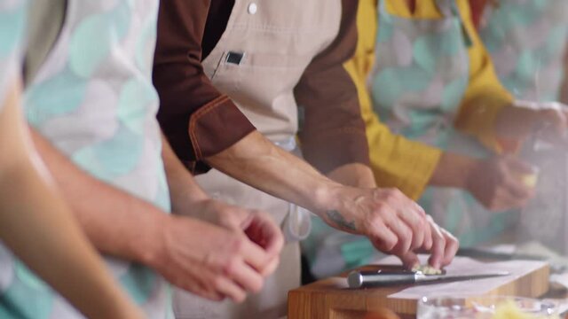 Tilt up shot of male chef and group of students smashing garlic cloves with knives while cooking together during culinary class
