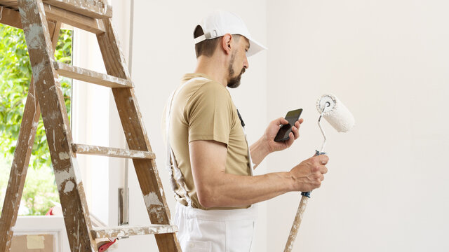 house painter man with mobile phone and paint roller, works the white home wall, a wooden ladder and green window as a background