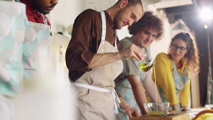Male chef in apron pouring olive oil into bowl and telling recipe to multiethnic students during cooking master class
