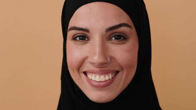 A close-up view of a happy arabian woman wearing a black hijab is opening her eyes and smiling to the camera standing isolated over a beige wall in the studio