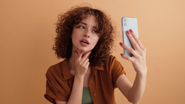 A smiling woman with curly hair is taking selfie photo using her smartphone standing isolated over a beige wall in the studio