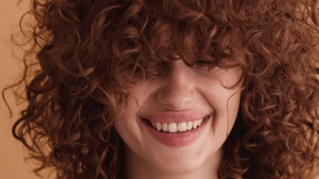 A close-up view of a curly woman is posing standing isolated over a beige wall in the studio
