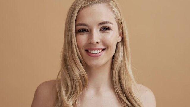 A beautiful blonde woman is posing to the camera while standing isolated over a beige wall in the studio