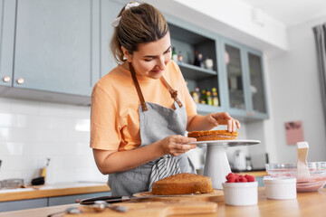 culinary, baking and cooking food concept - happy smiling young woman making layer cake on kitchen...