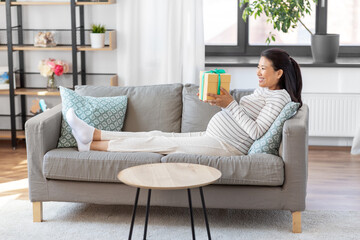 pregnancy, holidays and people concept - happy smiling pregnant asian woman with gift box sitting on sofa at home