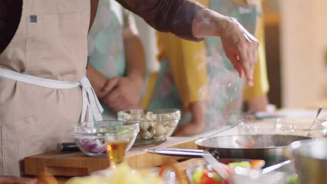 Midsection shot of unrecognizable male chef in apron adding spices to food on frying pan while cooking during culinary class