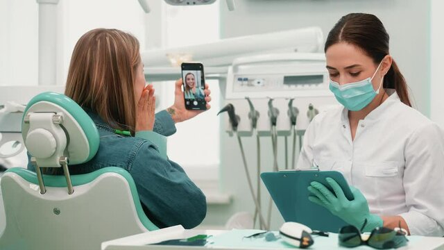 A dentist woman wearing a protective mask is making notes while her client is taking selfie photo sitting in a medical clinic at dental clinic