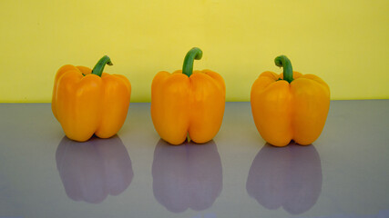 Three orange peppers on a yellow background stand upright on a gray table top. Shadows are reflected in the table top.