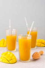 Freshly squeezed mango juice in glasses with a straw, ripe cut mango on a gray background