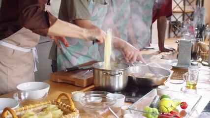 Professional chef in apron taking pasta from pot with tongues and putting it on frying pan while male student stirring during cooking master class